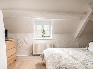 Quarto com teto inclinado, janela com planta, radiador, cómoda de madeira e cama com manta fofa.