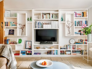 A living room with a flat-screen TV, bookshelves, a coffee table with fruit, and a clock on the wall. - 4 bedroom accommodation in Shilton (Shilton)