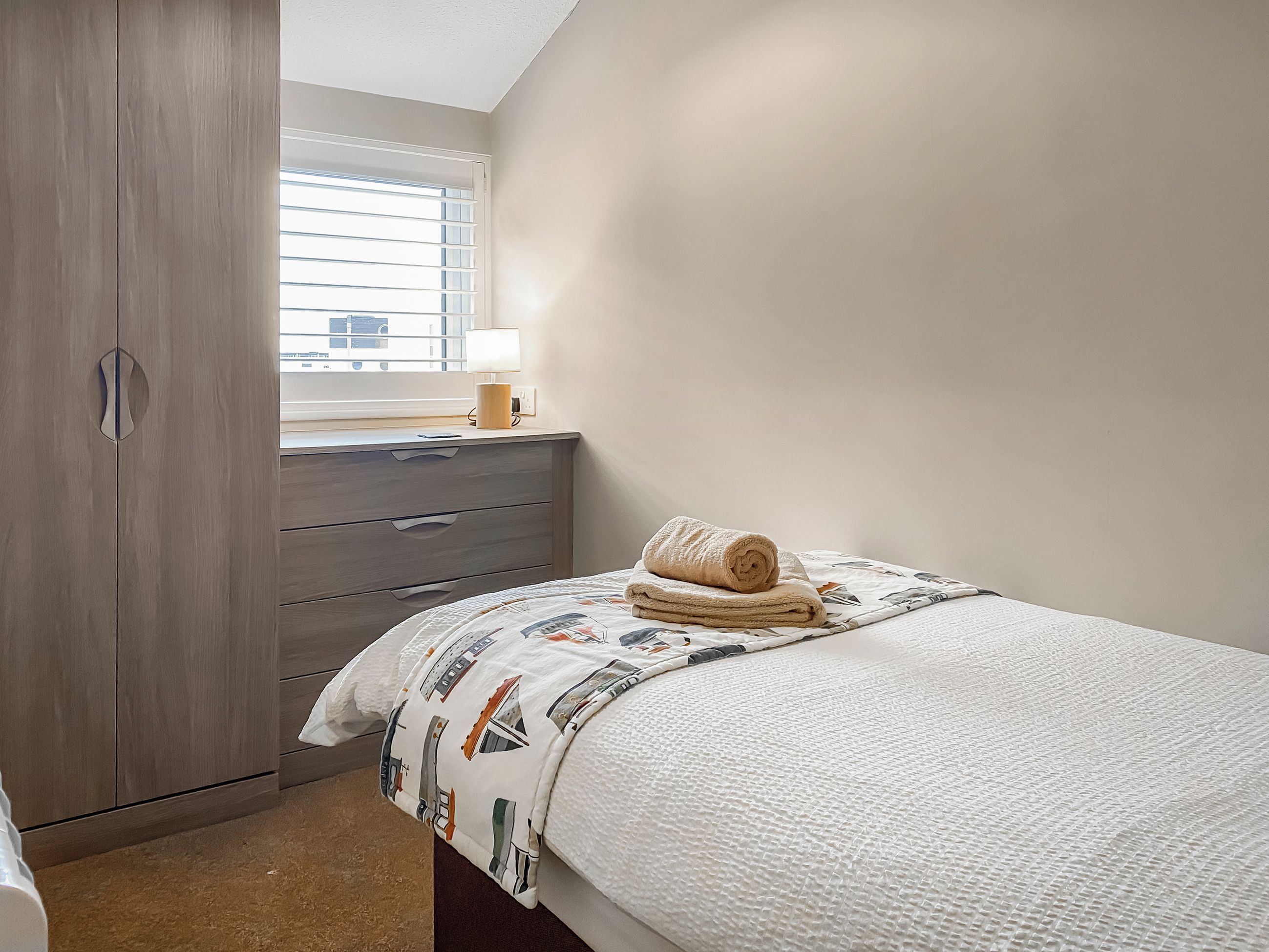 A bedroom with a bed, a bedside table with a lamp, a wardrobe, and a window with blinds.