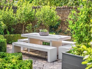 An outdoor patio with a white table and benches, surrounded by greenery and a stone wall. - 3 bedroom accommodation in Southrop (Southrop)