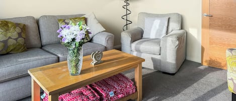 Un salon avec un canapé gris, un fauteuil gris, une table basse en bois et un vase rempli de fleurs.