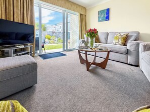 A modern living room with a flat-screen TV, a grey sofa, a coffee table with a vase of red flowers, and a glass door leading to a balcony. - Kingfisher 9 - UKC9969 (Christchurch)