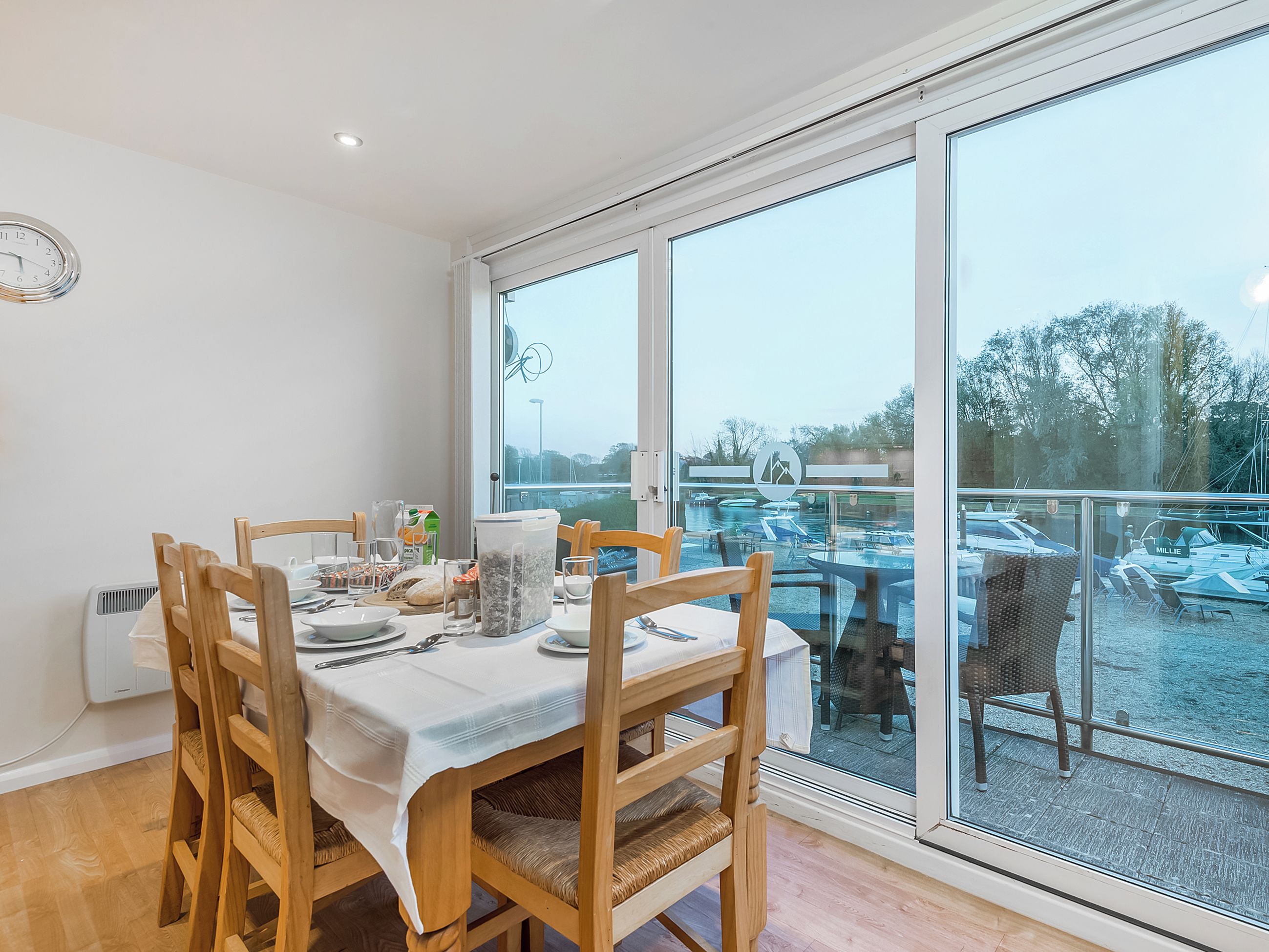 Comedor con una mesa dispuesta para cuatro, sillas de madera y vista a un puerto deportivo a través de amplias ventanas.