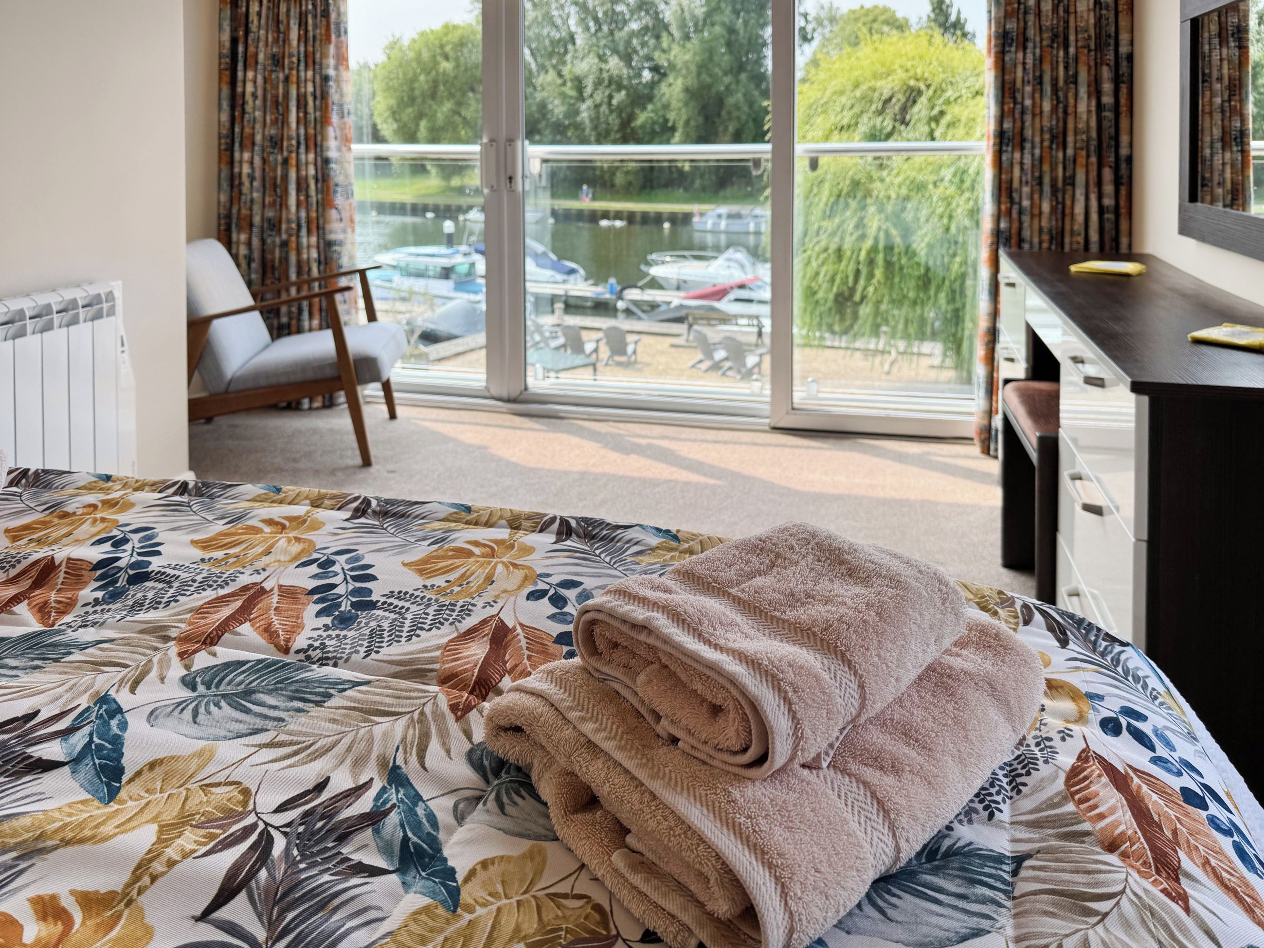 A hotel room with a bed, a chair, a desk, and a view of a body of water through large windows.