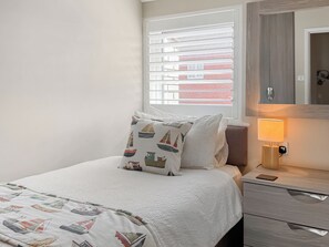 Ein ordentlich bezogenes Bett mit Kopfkissen, die ein Segelbootmuster zeigen, eine Nachttischlampe und ein Fenster mit Jalousien.