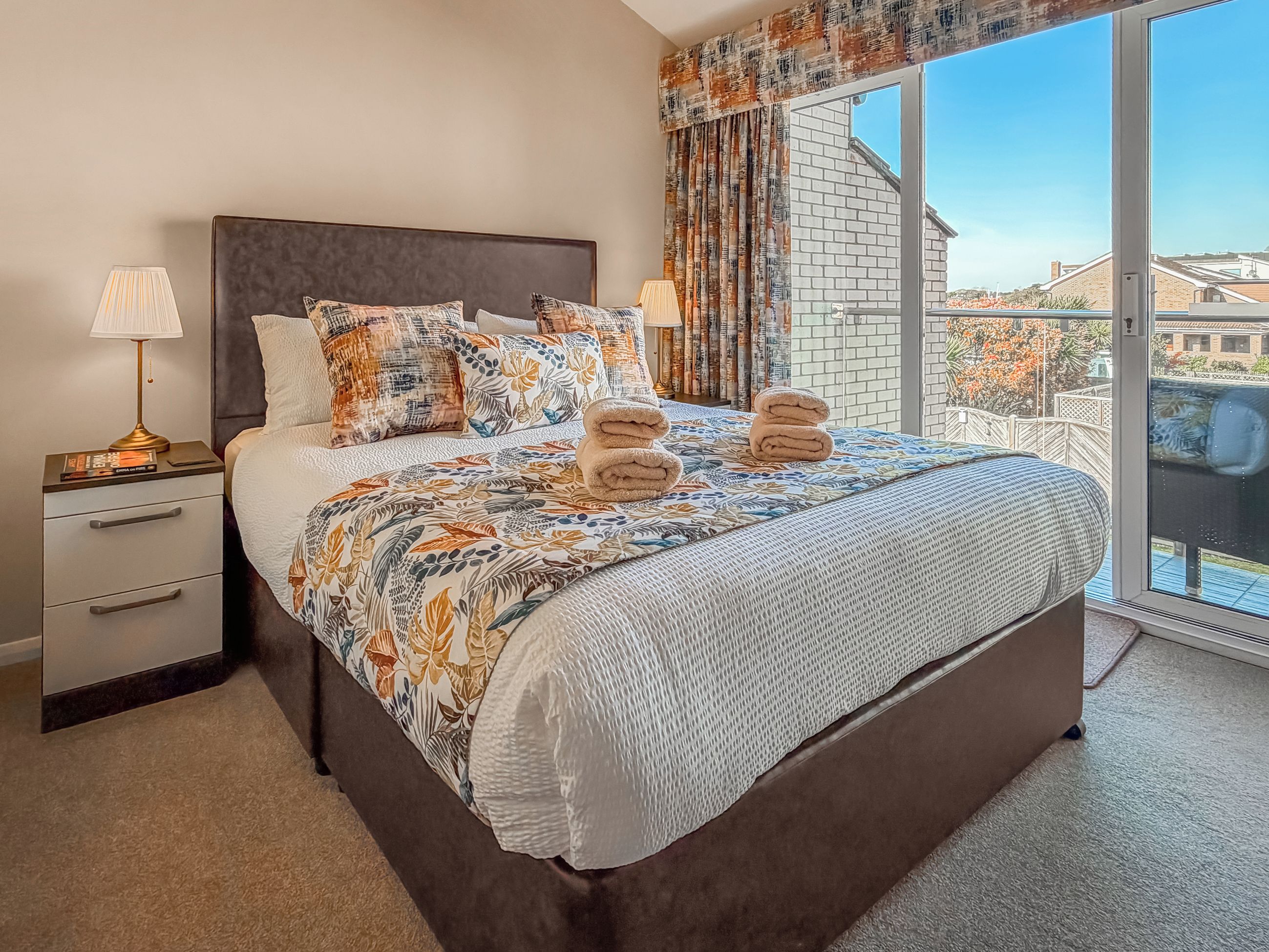 Une chambre d’hôtel avec un grand lit, deux tables de chevet, une fenêtre donnant sur des immeubles et un balcon.