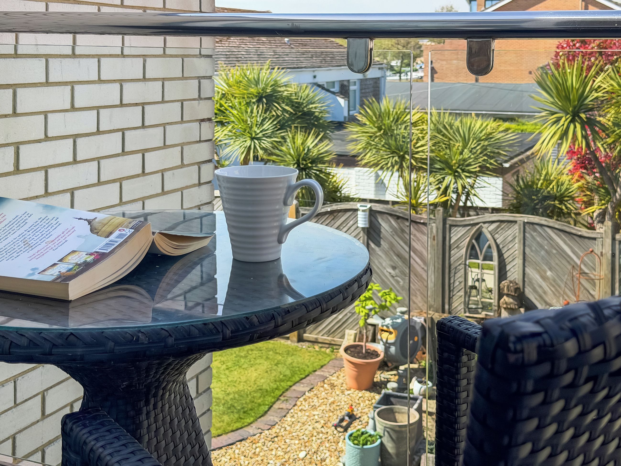 Eine Terrasse mit einem Glastisch, einer weißen Tasse und einem Buch. Im Hintergrund sind eine Backsteinmauer und ein Holzzaun zu sehen.