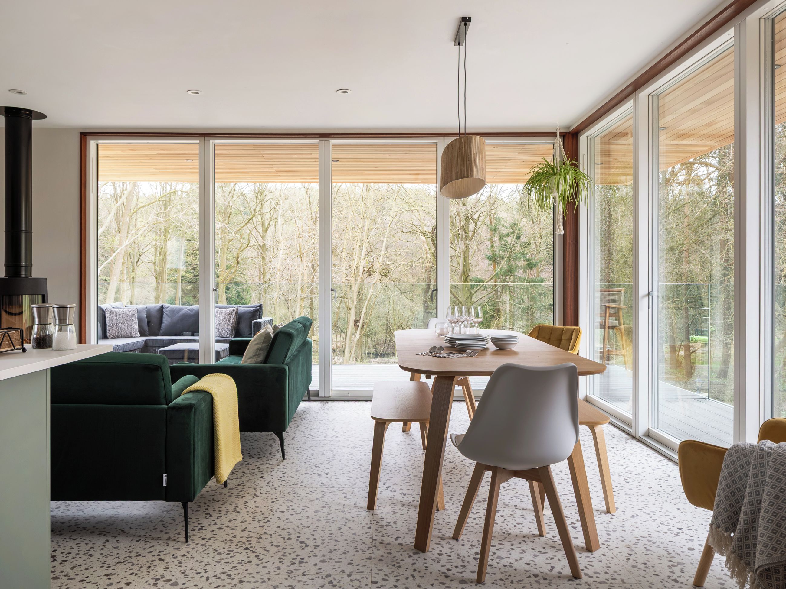 A modern interior with a dining area, a sofa set, and large windows offering a view of trees.