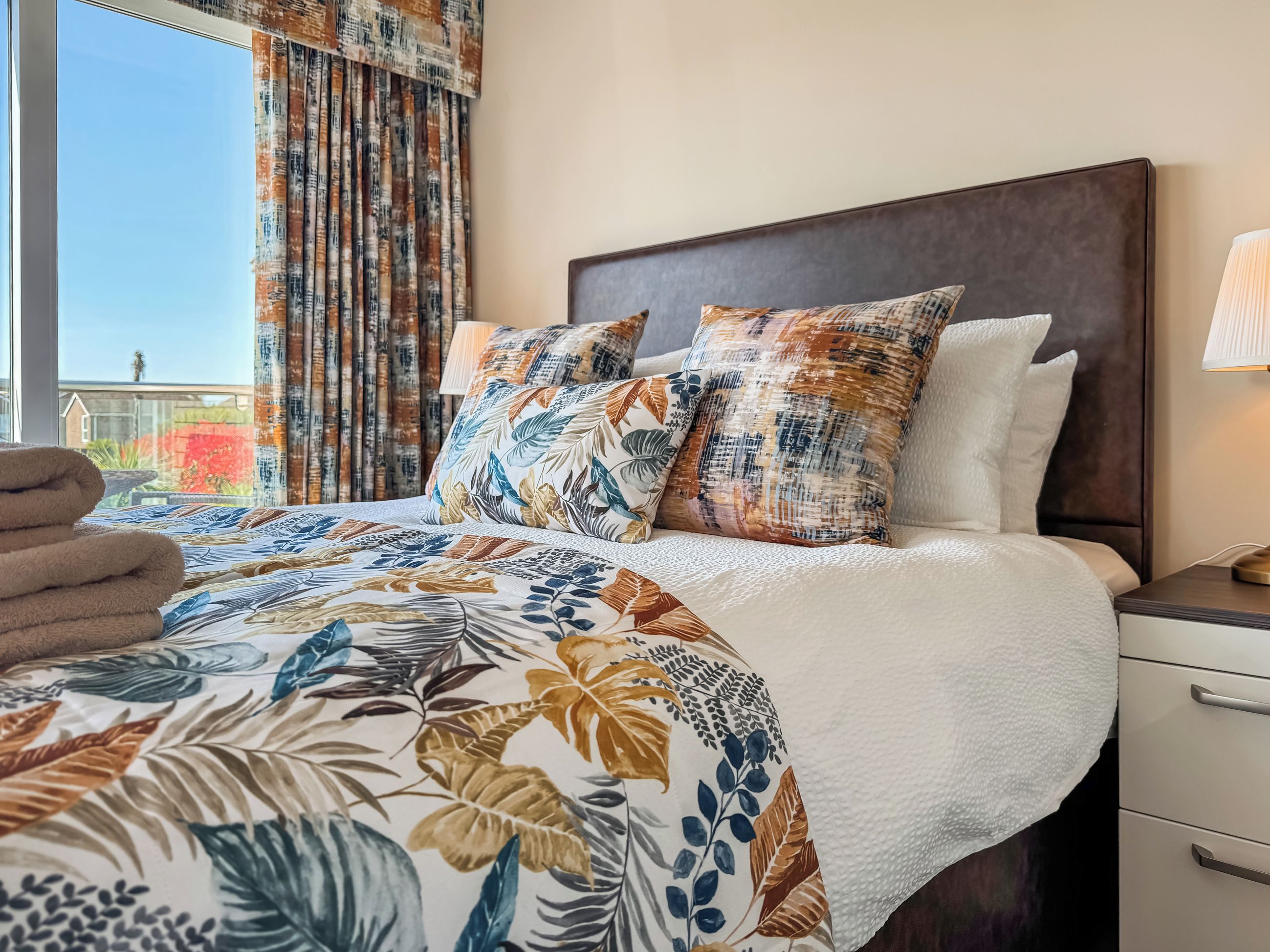 A hotel room with a bed, a nightstand, a lamp, and a view of the outside.