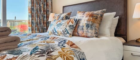 A hotel room with a bed, a nightstand, a lamp, and a view of the outside.