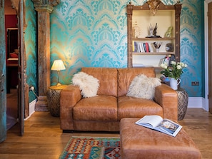 Un canapé en cuir marron avec deux coussins, une petite table d’appoint avec une lampe, une bibliothèque remplie de livres et d’objets décoratifs, et un tapis à motifs sur un plancher en bois.