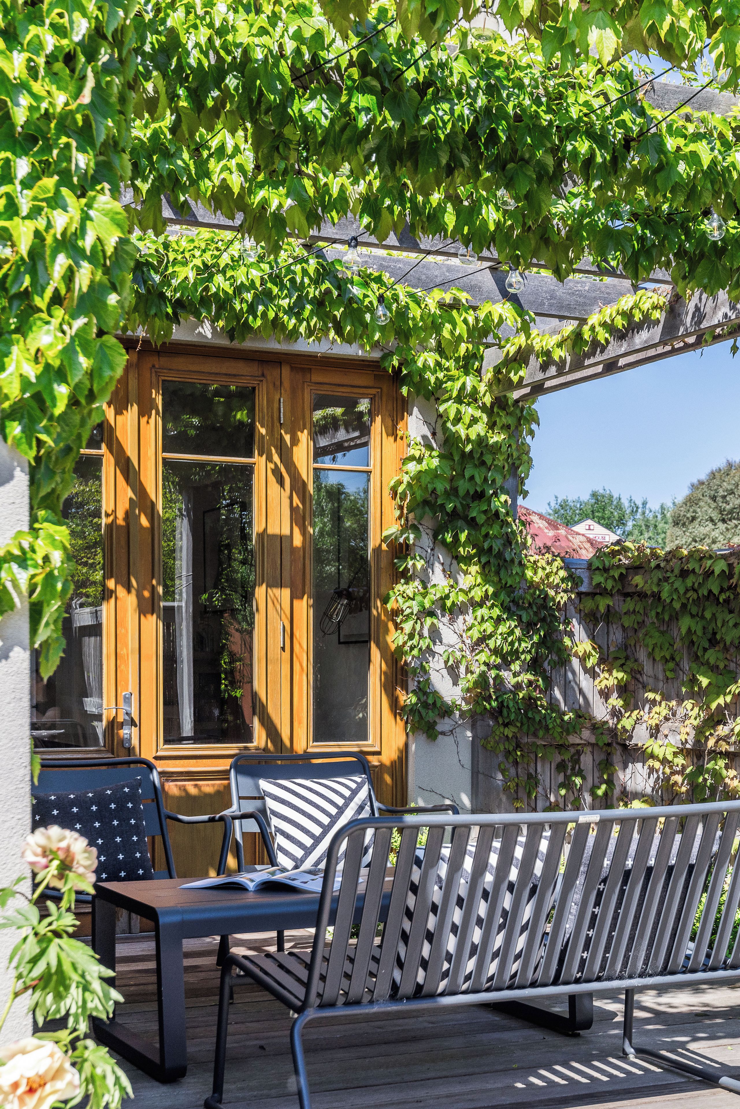 Una pérgola de madera con puerta de cristal, un banco de madera, y una mesa con sillas.