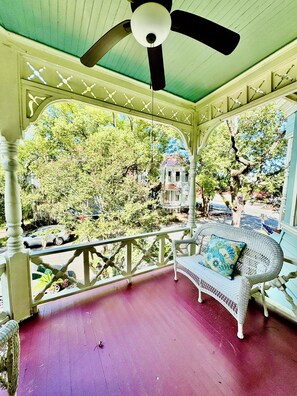 Eine überdachte Veranda mit Deckenventilator, ein Korbsessel mit Kissen und Blick auf Bäume sowie ein Haus im Hintergrund.