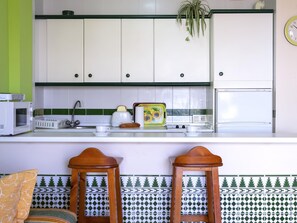 A kitchen with white cabinets, a microwave, a toaster, and a sink. There are two wooden stools and a patterned tile backsplash. - Sunstay Relax 48 Rio Marina Nerja (Nerja)