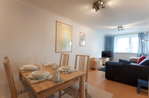 Un comedor con una mesa de madera dispuesta para cuatro personas, un sofá, un televisor y un ventanal con cortinas.