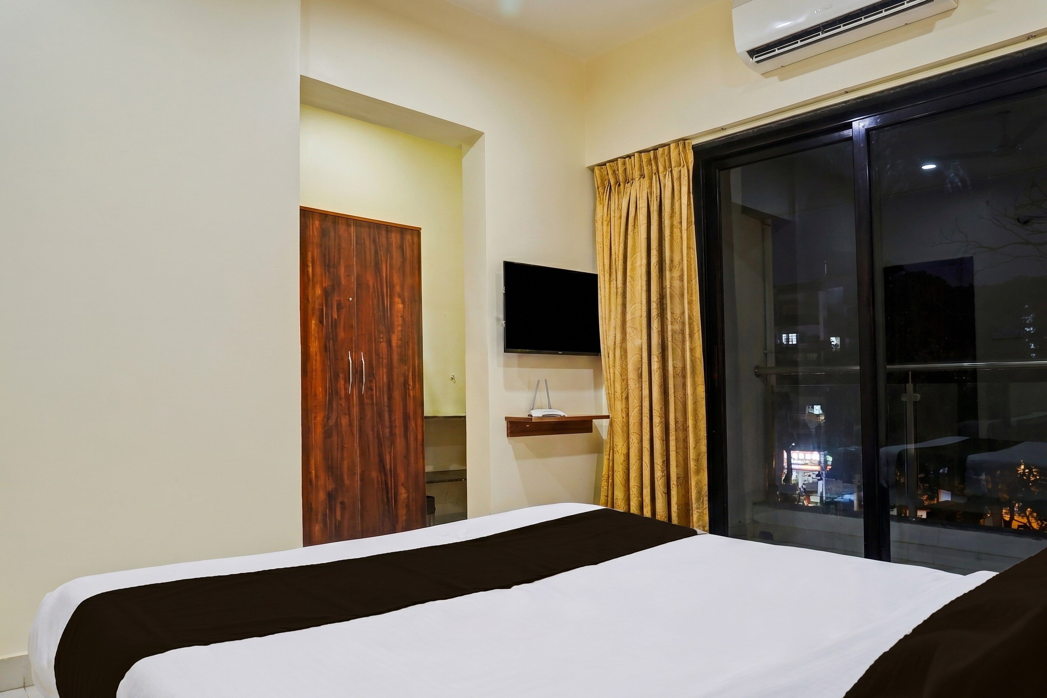 A hotel room with a bed, a TV, a window with curtains, and an air conditioning unit.