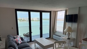 Een woonkamer met een bank, een tafel en een stoel. Er is een balkon met uitzicht op het water en gebouwen.
