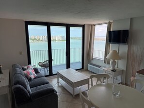 Un salon avec un canapé, une table et une chaise. Il y a un balcon d’où l’on peut admirer la vue sur l’eau et les bâtiments.