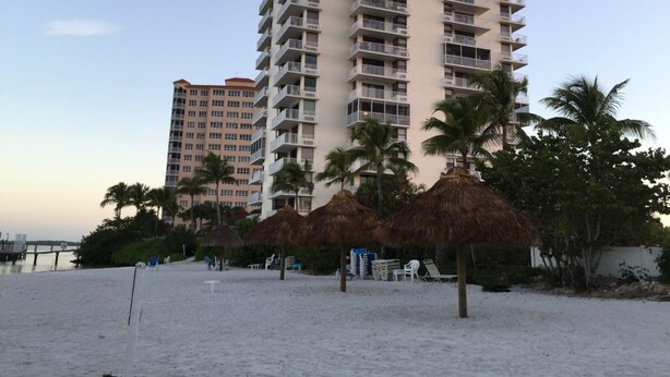 Een hotel aan het strand met meerdere balkons, palmbomen en rieten parasols.