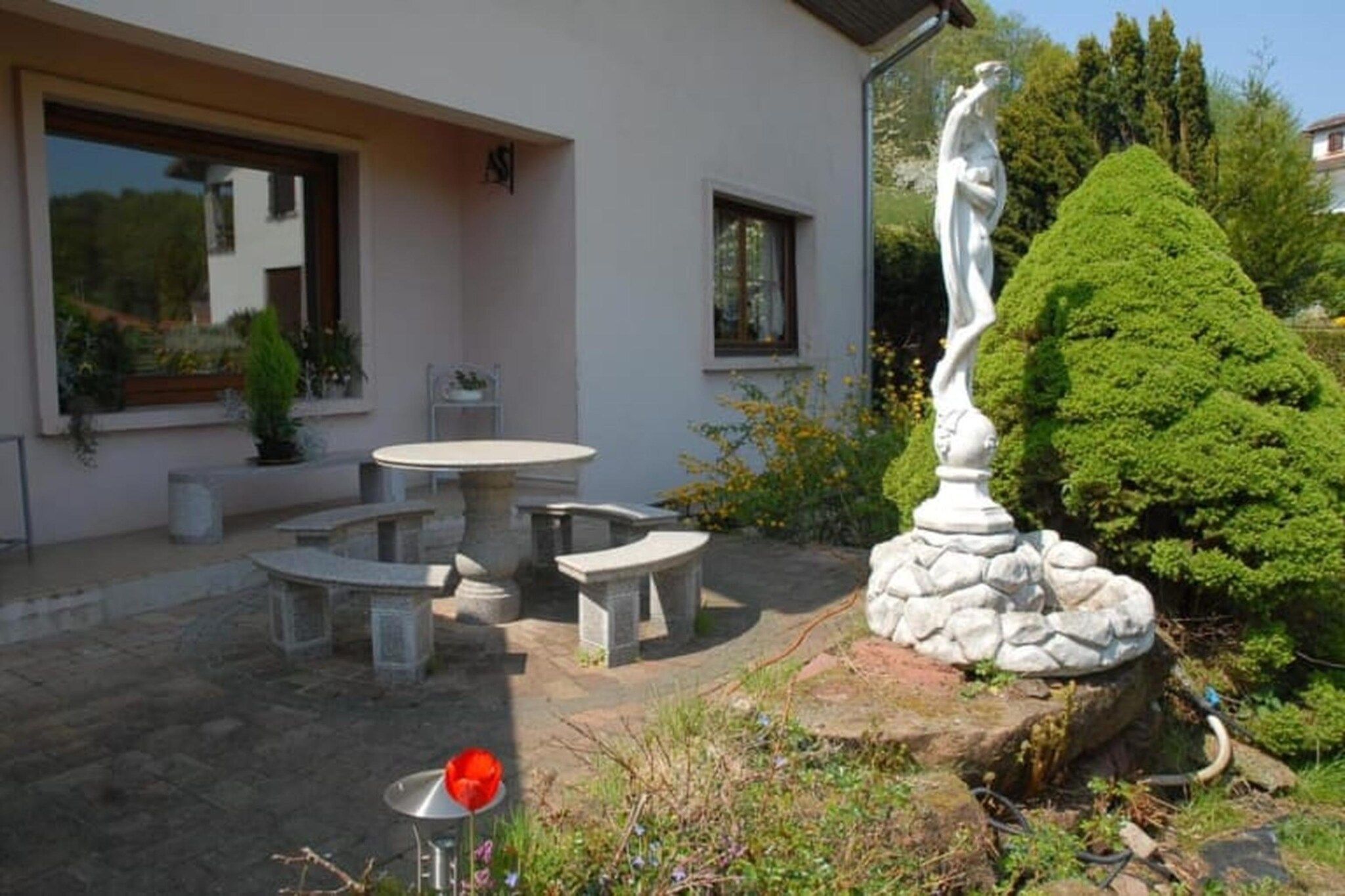 A patio with a stone table and benches, a statue, and a well-maintained garden with a hedge.