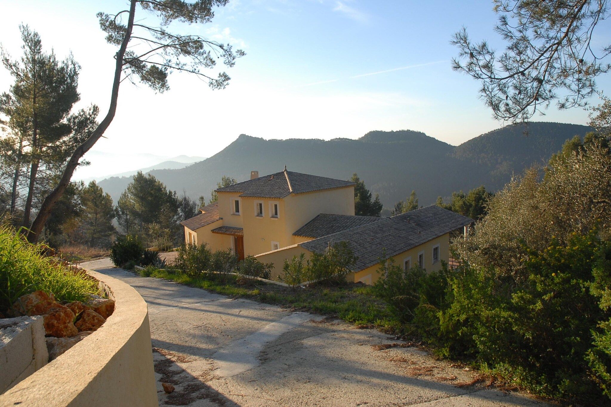 A two-story building with a sloped roof, surrounded by greenery and mountains in the background.