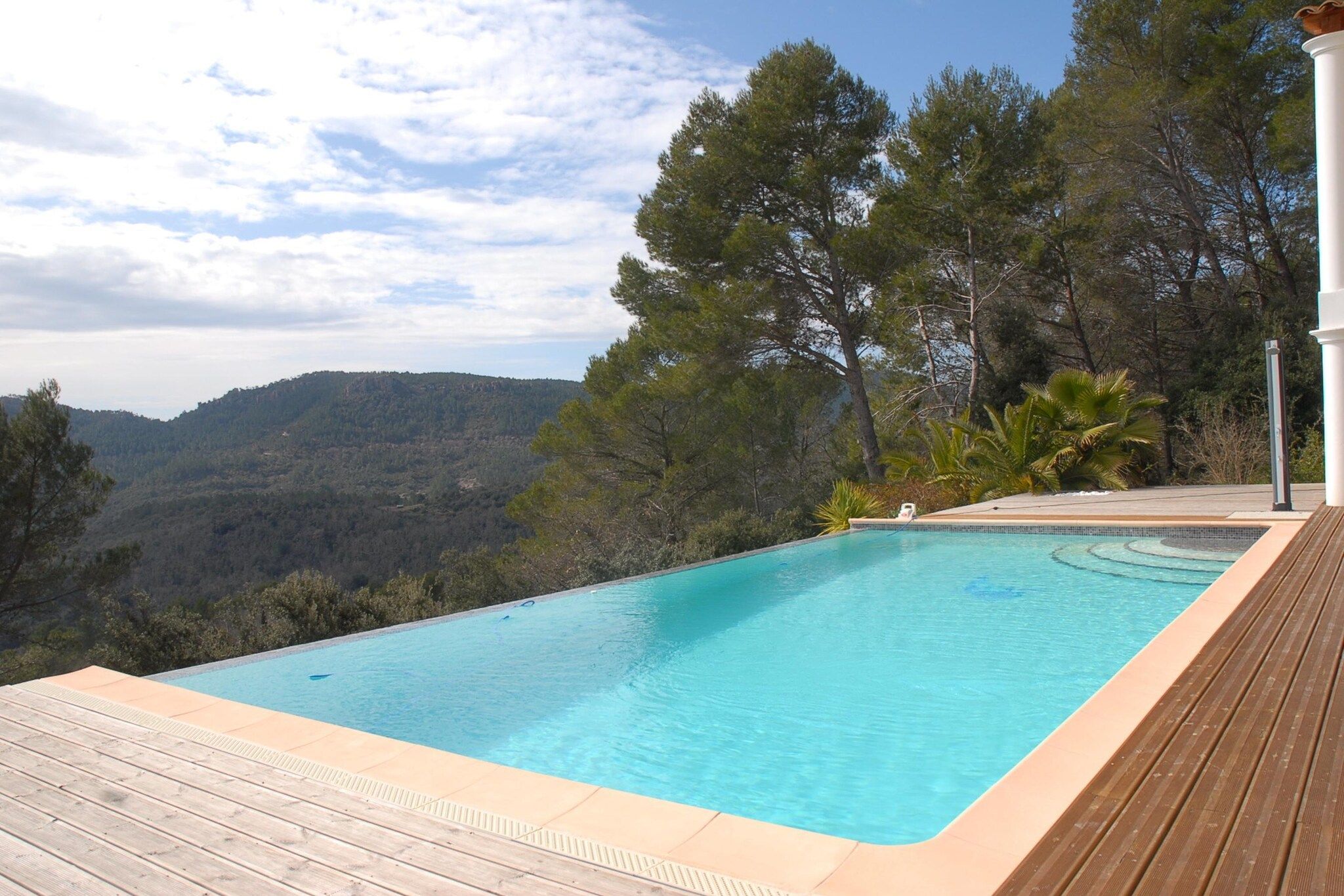 A swimming pool with a wooden deck, surrounded by trees and a mountainous landscape.