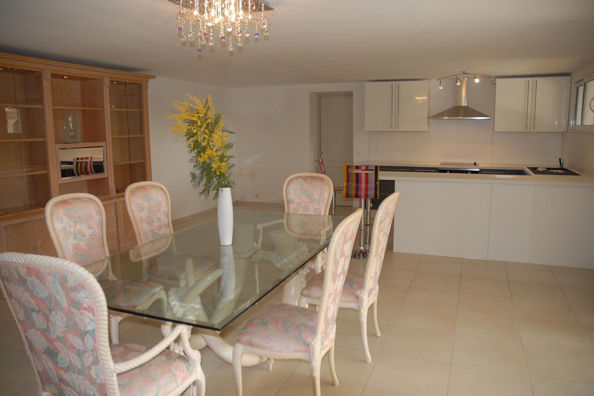 A dining area with a glass table surrounded by floral chairs, a vase with yellow flowers, and a kitchen in the background.