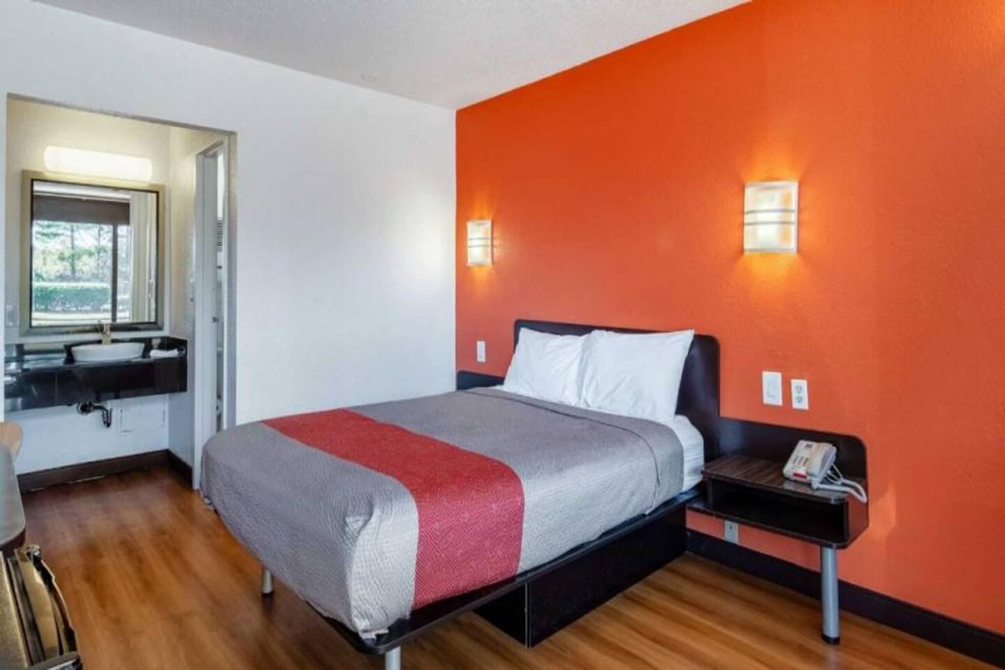 A hotel room with a bed, a nightstand, a wall-mounted phone, and a window with blinds.