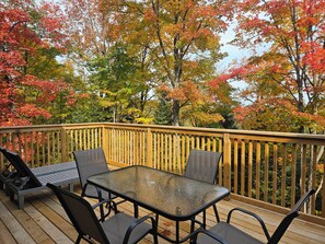 A wooden deck with a glass table and chairs, surrounded by trees with autumn foliage. - Beautiful Muskoka Cottage with Games, BBQ & Views (Huntsville)