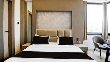 Habitación de hotel con una cama grande, una mesita de noche con lámpara, una silla y vista a la ciudad desde la ventana.