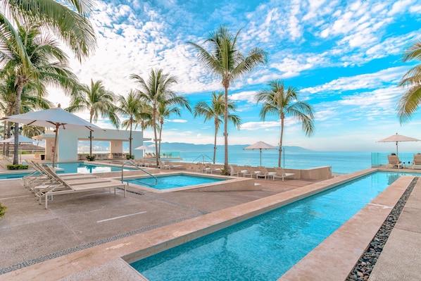 A pool area with lounge chairs and umbrellas, palm trees, and a view of the ocean. - Stunning Oceanfront 3BR, Rooftop + 7 pools in PV! 3BD Condo for rent in North Ho (Puerto Vallarta)