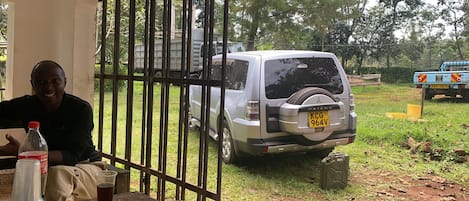 Una persona sentada a una mesa con una botella y un vaso, un vehículo estacionado y una verja al fondo.
