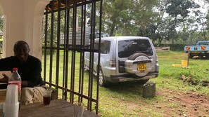 A person seated at a table with a bottle and a glass, a parked vehicle, and a gate in the background.