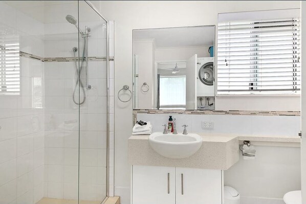 A modern bathroom with a glass shower enclosure, a sink with a mirror above it, and a toilet. - Mooloolaba Magic 2 min to the Beach, Wharf & Rooftop Living at Your Doorstep (Mooloolaba)