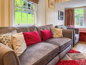 Ein Wohnzimmer mit einem braunen Sofa, roten und gemusterten Kissen, einem roten Teppich und einem Fenster mit Blick auf Grünflächen.