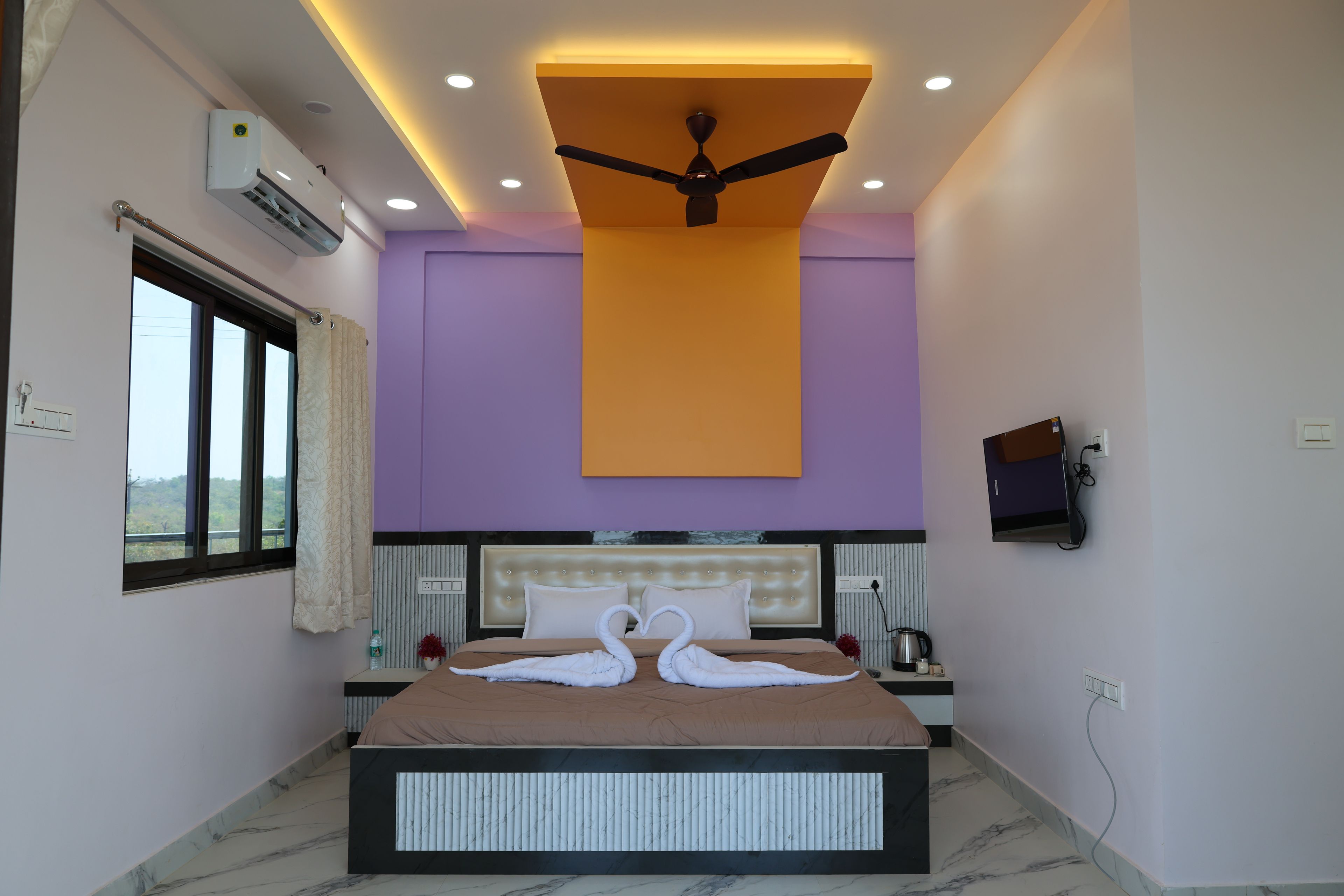 Habitación de hotel con cama, ventilador de techo, aire acondicionado, televisor montado en la pared y ventana con vistas.