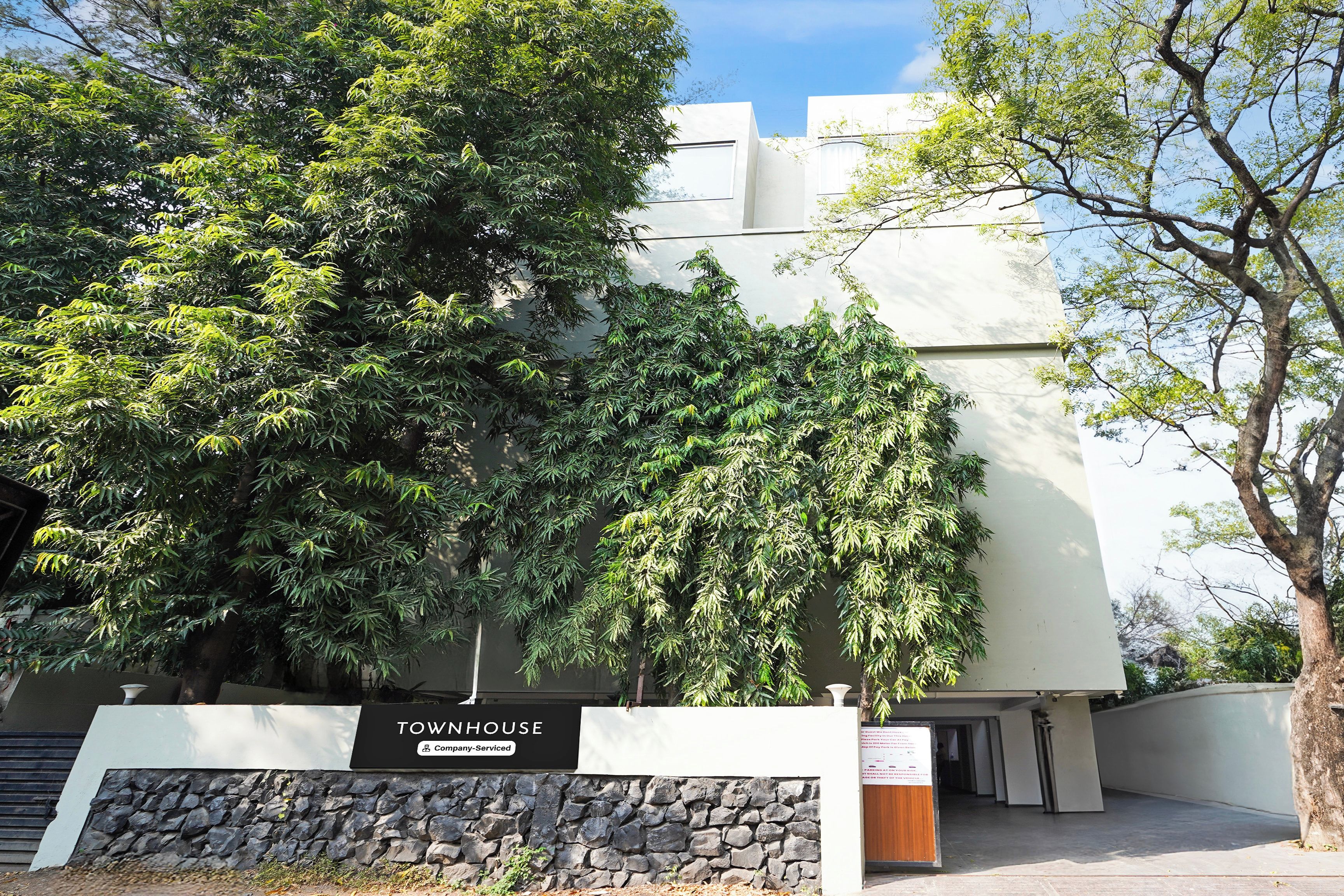 Uma casa geminada com entrada em parede de pedra, placa indicando serviço corporativo e uma grande árvore em frente.
