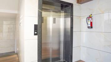 Un ascensor moderno con acabado metálico, un extintor de incendios montado en la pared y un suelo de madera.