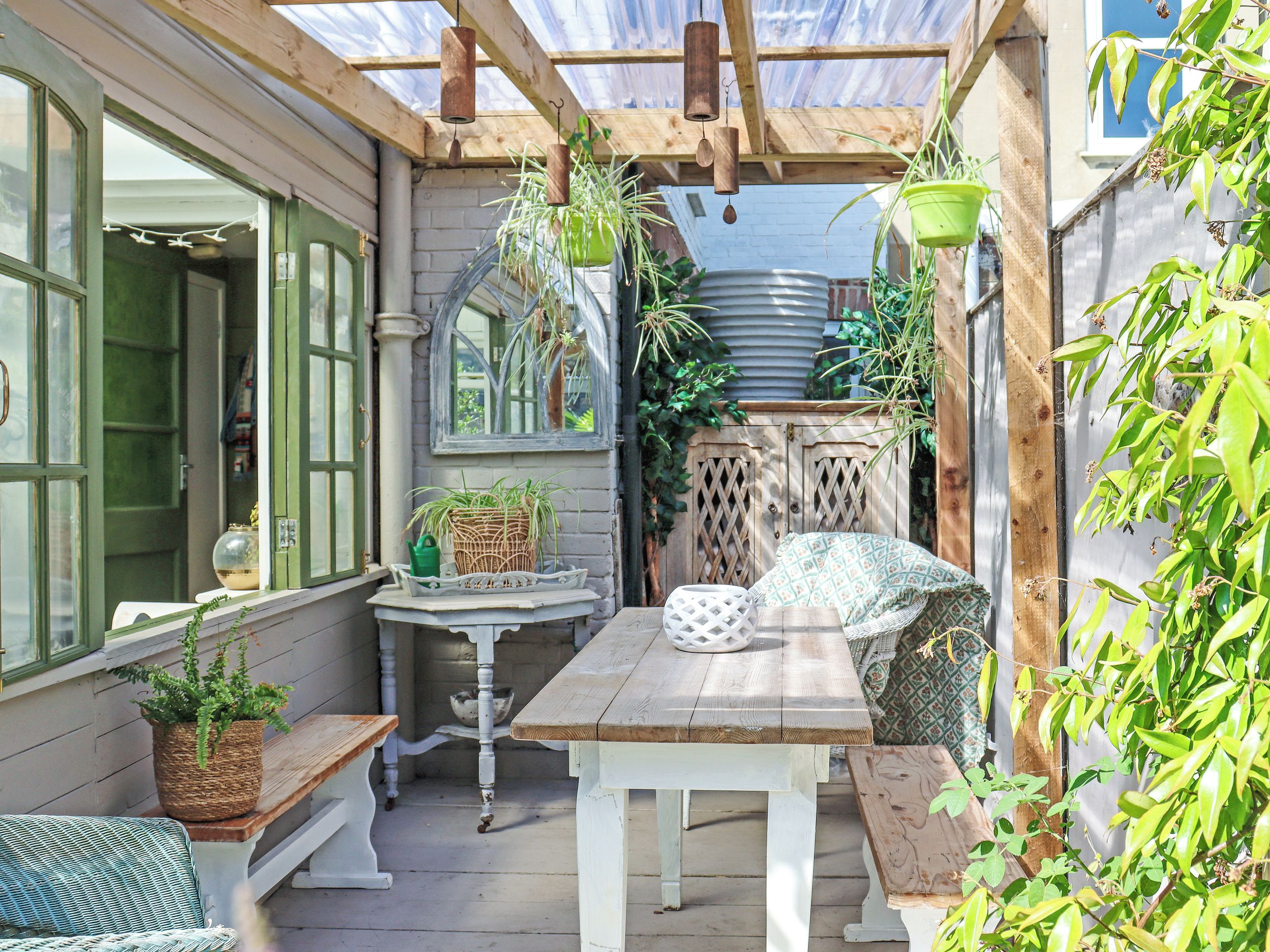 Een overdekt terras met een houten tafel en bankje, omringd door potplanten en een hanglamp.