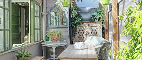 Une terrasse couverte avec une table et un banc en bois, entourée de plantes en pot et agrémentée d’un luminaire suspendu.