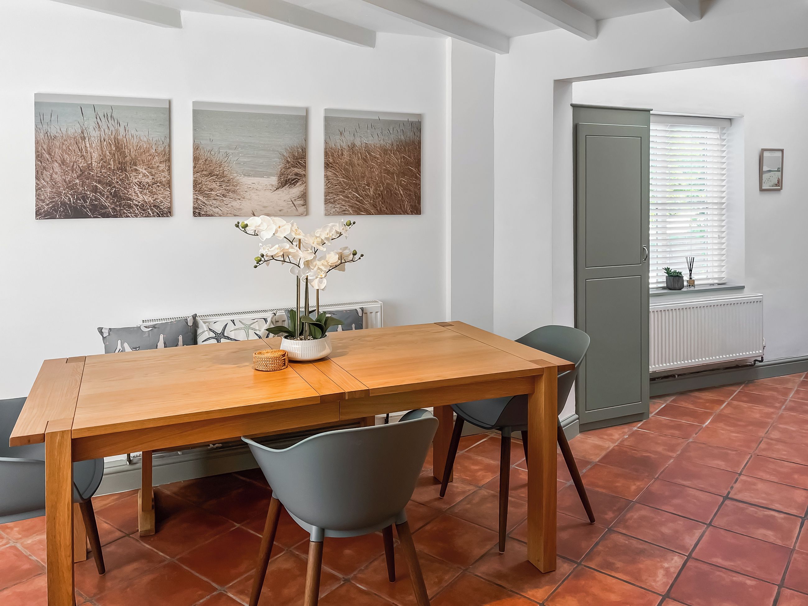 A dining area with a wooden table, grey chairs, and a vase with white flowers. There are three framed pictures on the wall.