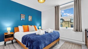 A bedroom with a bed, nightstands, a window with curtains, and a view of buildings outside. - A terraced cottage on Bennett Street, in the heart of Buxton. (Buxton)