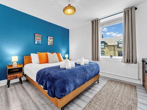 A bedroom with a bed, nightstands, a window with curtains, and a view of buildings outside. - A terraced cottage on Bennett Street, in the heart of Buxton. (Buxton)