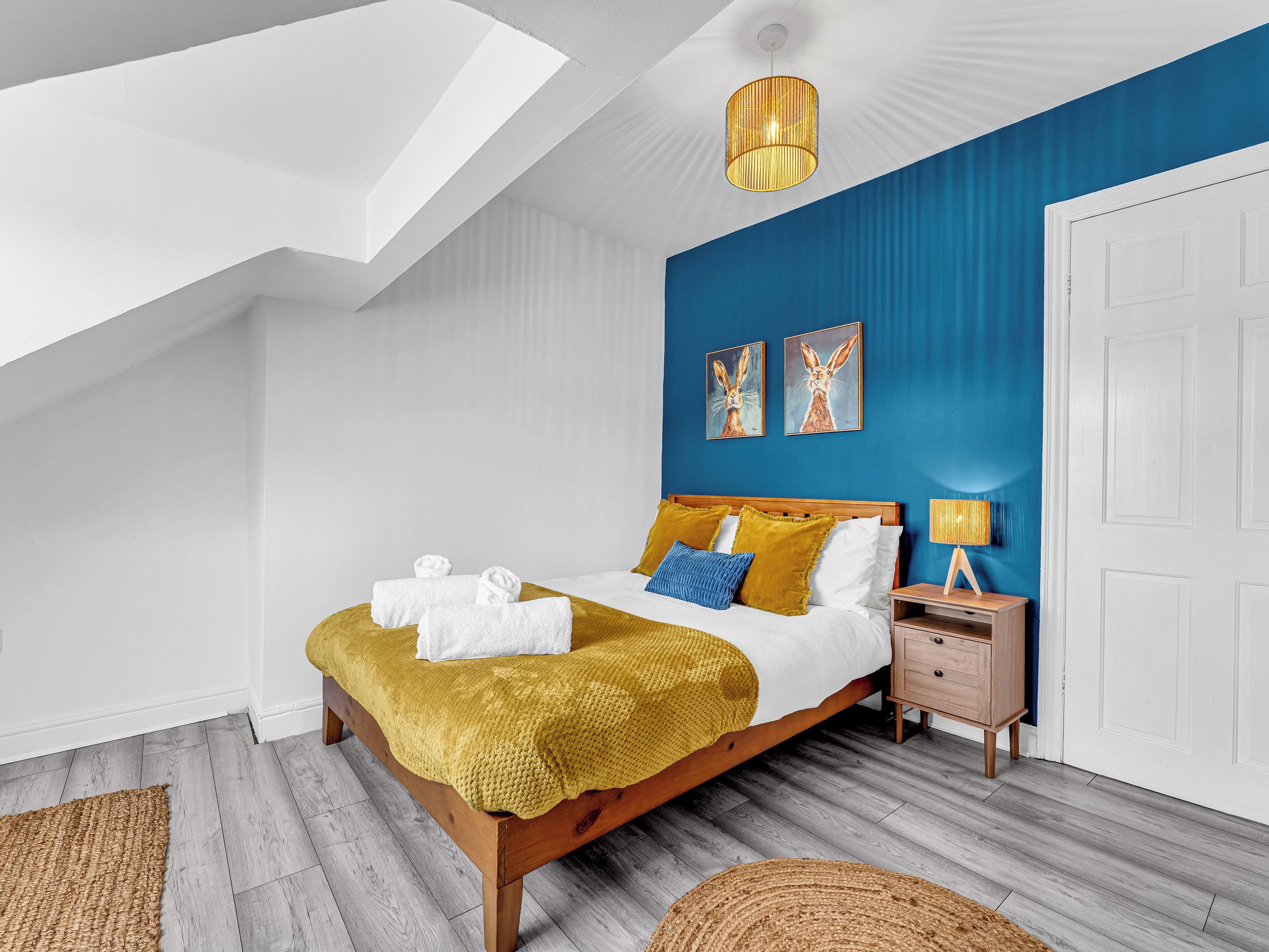 Une chambre moderne avec un plafond en pente, un lit agrémenté d’un jeté jaune, des serviettes blanches et deux tableaux accrochés au mur.