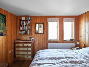 Ein Schlafzimmer mit Holzwänden, einem Bett, einem Schminktisch, einem Bücherregal und zwei mit Vorhängen versehenen Fenstern.