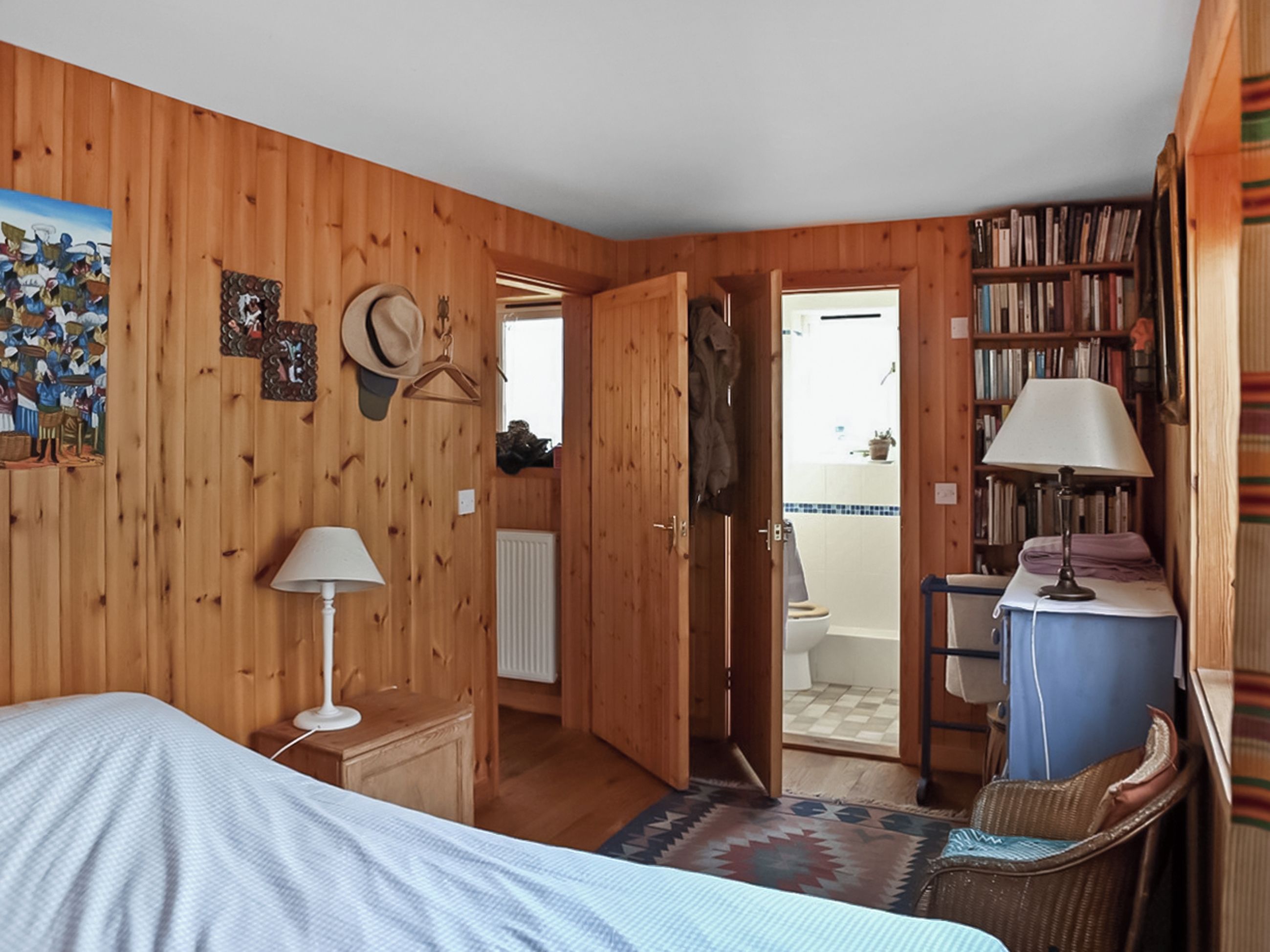 A wooden room with a bed, a desk, a chair, a hanging hat, a lamp, and a bookshelf.