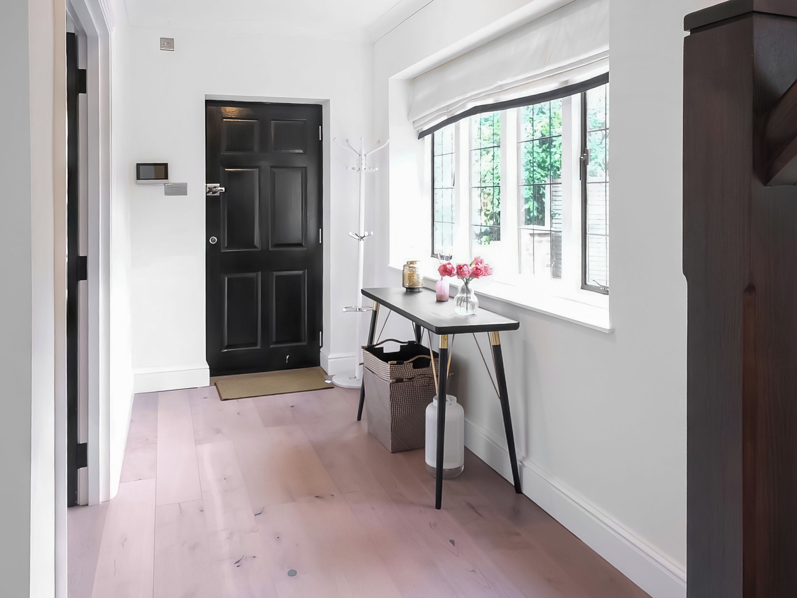 Un couloir avec une porte noire, un mur blanc, une fenêtre donnant sur un paysage verdoyant et une petite table sur laquelle est posé un vase de fleurs.