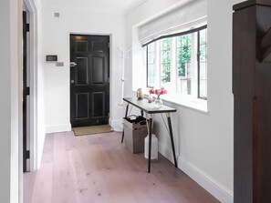 Un couloir avec une porte noire, un mur blanc, une fenêtre donnant sur un paysage verdoyant et une petite table sur laquelle est posé un vase de fleurs.