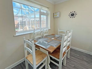 A dining area with a table and chairs, a window with a view, and a wall clock. - 3 bedroom accommodation in Hogsthorpe (Hogsthorpe)