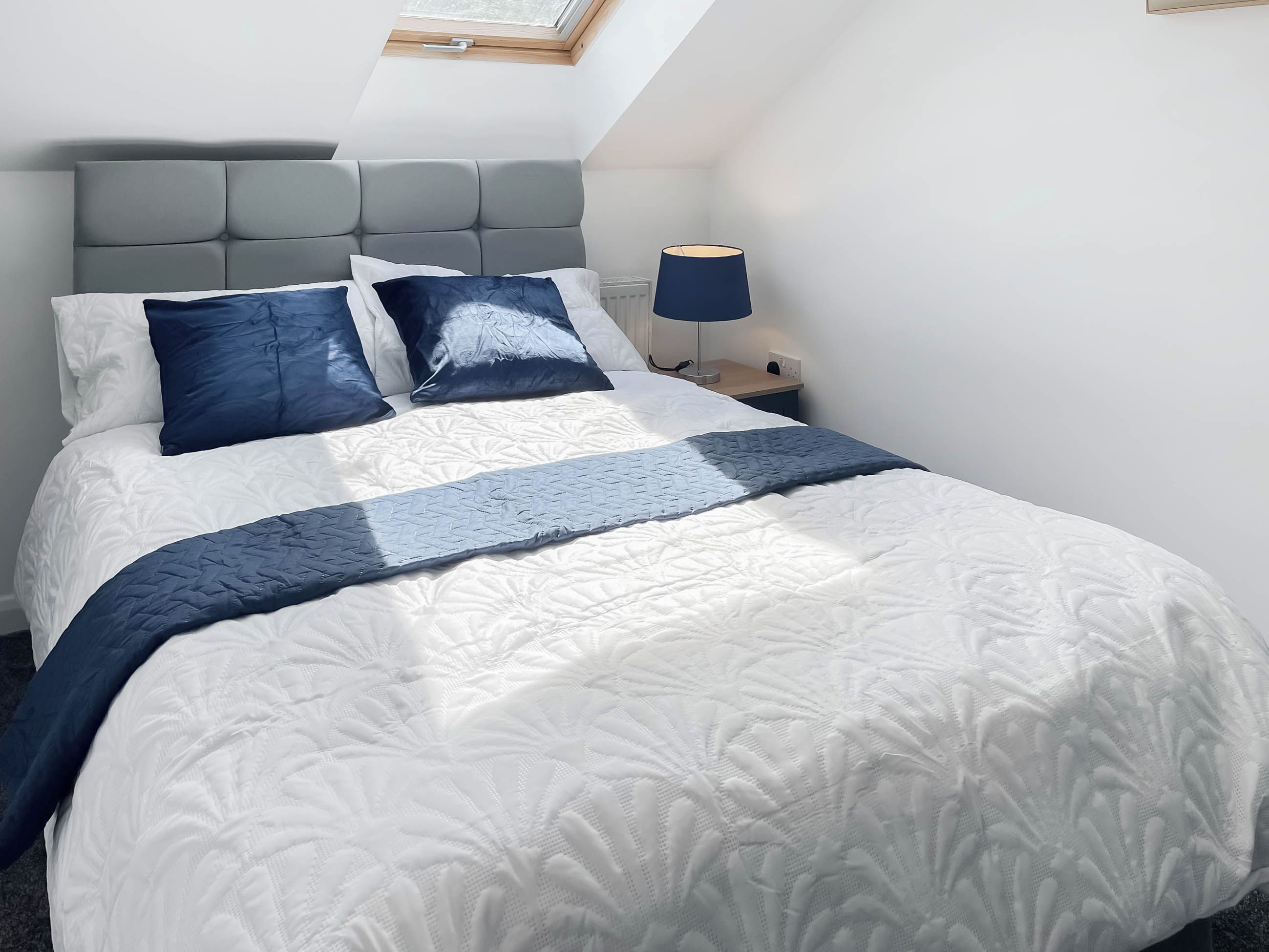 A neatly made bed with a headboard, a bedside lamp, and a window above the bed.